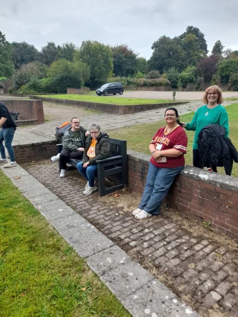 A small group of walkers sitting outside on a stone bench area near a green lawn. Two individuals are seated on a bench, while another stands beside them, smiling. It is a relaxed moment after the walk with greenery and trees in the background.