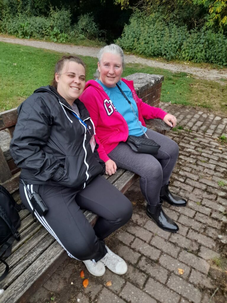 Two women seated on a wooden bench, smiling at the camera. Both are wearing comfortable casual clothes, suggesting they are taking a break after the walk in a relaxed outdoor setting.