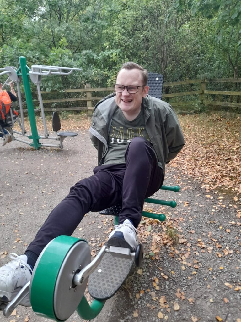 A man smiling and sitting on an outdoor exercise machine in a park. He appears to be having fun as he uses the equipment. The surrounding area is wooded, with leaves scattered on the ground.