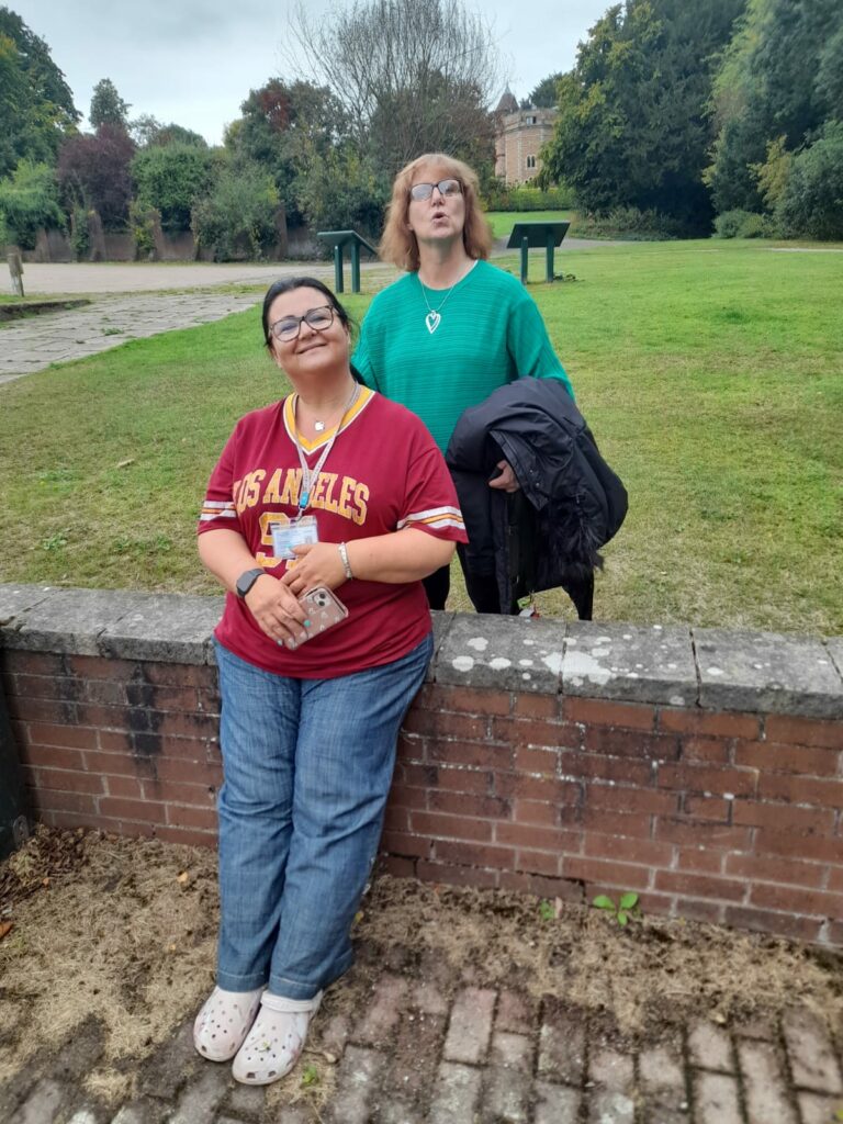 Two women are smiling as they lean against a brick wall outdoors. One is standing, and the other is seated, both appearing happy and relaxed. Behind them is a green, open area with trees and bushes.