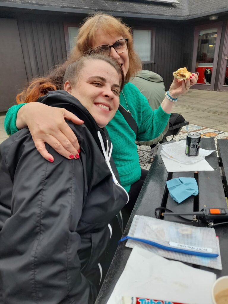 Two women sitting at an outdoor café table, one hugging the other. They are smiling, with food and drinks, including a sausage roll and a coffee, on the table in front of them. Both appear to be enjoying a friendly moment post-walk.