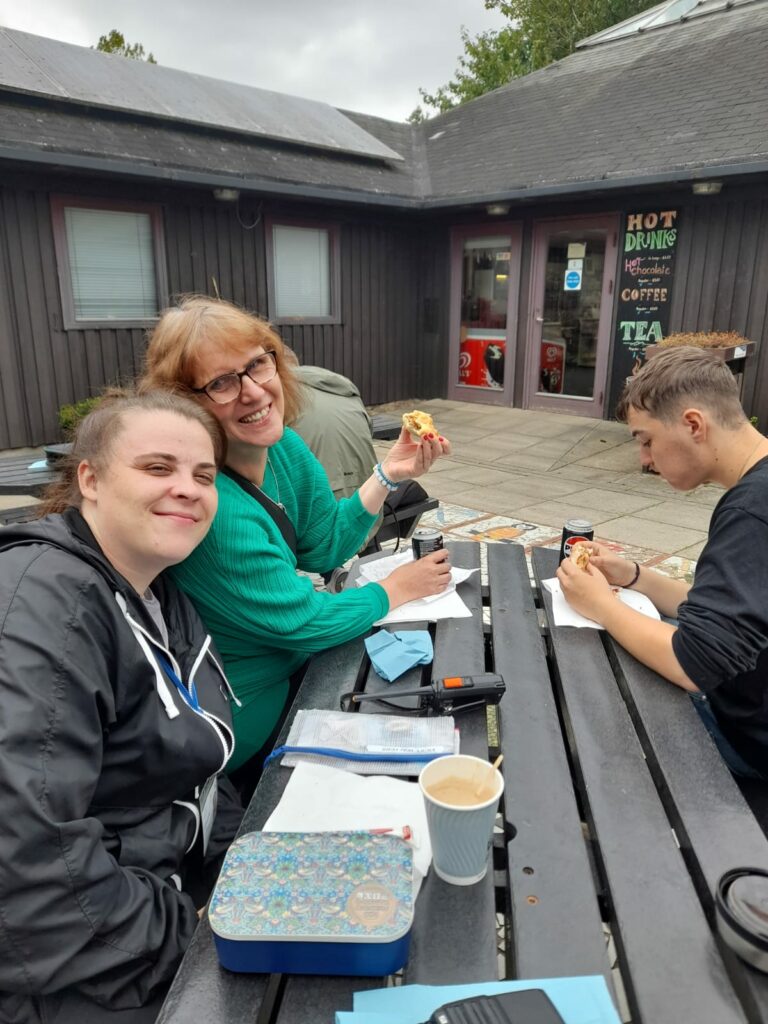 A group of walkers sitting at a picnic table outside a café, enjoying drinks and snacks. The table has cups, drinks, and sandwiches, and the people are smiling and engaged in conversation, with a cosy outdoor seating area in the background.