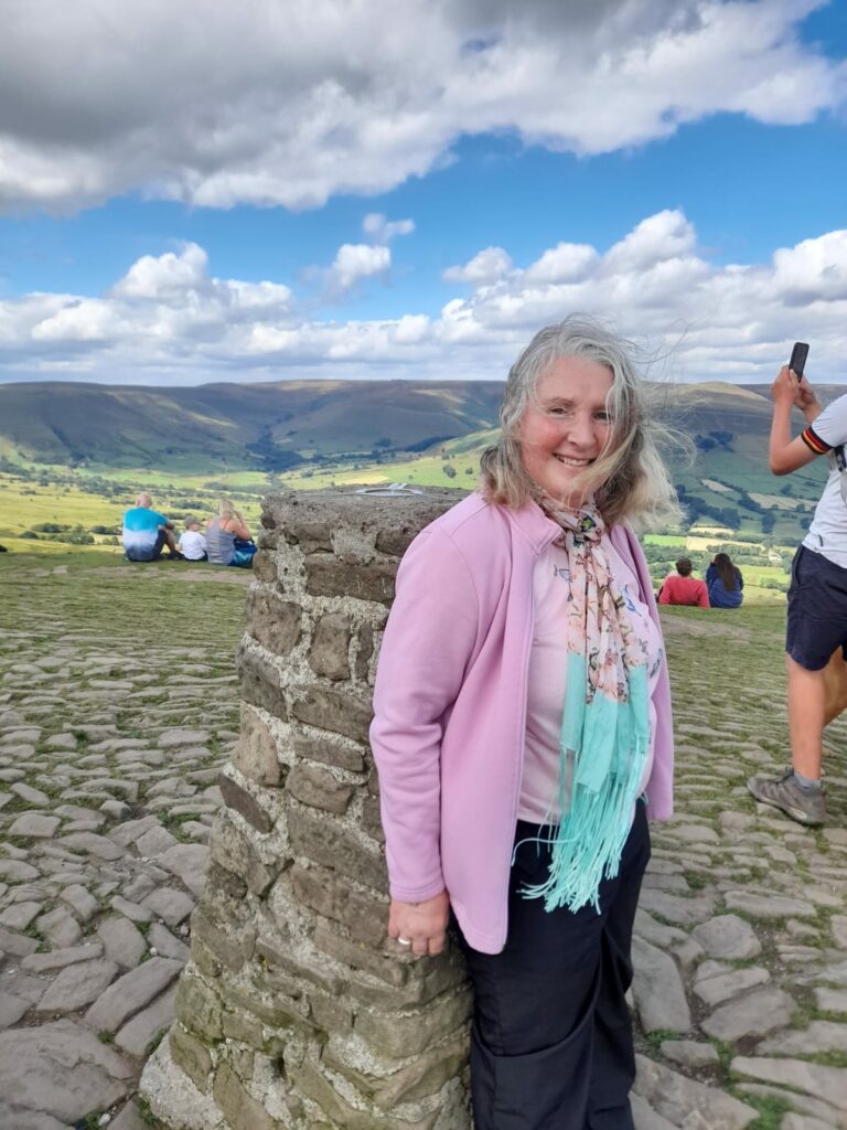 A woman standing and smiling next to a stone pillar at the summit of Mam Tor, with the stunning landscape of rolling green hills and valleys in the background under a partly cloudy sky. She is wearing a light pink jacket, a butterfly-patterned shirt, and a pastel scarf, with her hair blowing slightly in the wind. Other visitors are sitting on the grass in the background, admiring the view.