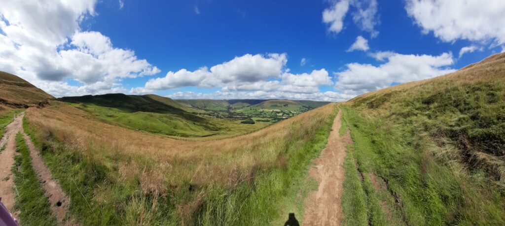 A wide, panoramic view of the landscape surrounding Mam Tor in the Peak District. The scene shows a dirt walking path winding through grassy hills, with expansive, rolling green hills and valleys in the background. The sky is bright blue with fluffy white clouds, creating a beautiful contrast with the natural terrain. The view captures the peaceful, open countryside, offering a sense of vastness and tranquility.