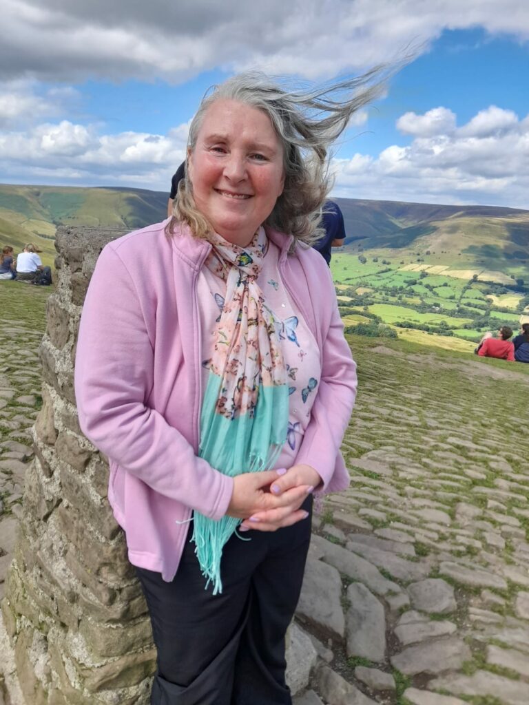 A woman standing and smiling near the summit of Mam Tor with a scenic landscape of green hills and valleys in the background. She is wearing a light pink jacket, a butterfly-patterned shirt, and a pastel scarf, with her hair being gently blown by the wind. The ground is made of cobblestone, and other people can be seen sitting and admiring the view in the distance under a partly cloudy sky.