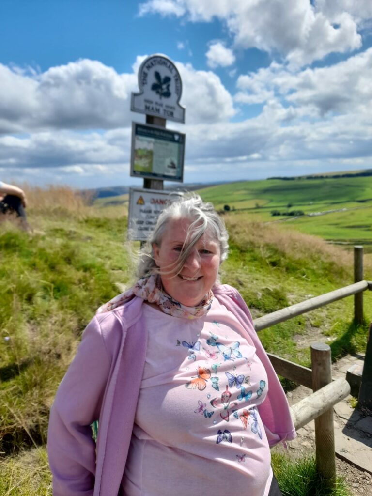 A woman smiling while standing outdoors near the Mam Tor signpost, with rolling green hills in the background under a partly cloudy sky. She is wearing a light pink jacket over a butterfly-patterned shirt and a pastel scarf, with wind blowing her hair. A wooden fence and grassy area are visible behind her.