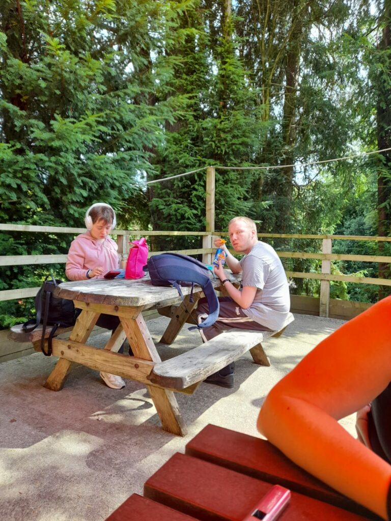 Two people sitting at a wooden picnic table outdoors, surrounded by tall trees. One person, wearing headphones and a pink hoodie, is looking down, possibly at a phone or tablet. The other person, with blonde hair and wearing a grey t-shirt, is eating a snack and looking towards the camera. Backpacks and a pink water bottle are placed on the table. The area is fenced, and sunlight filters through the trees, casting light and shadow across the scene.