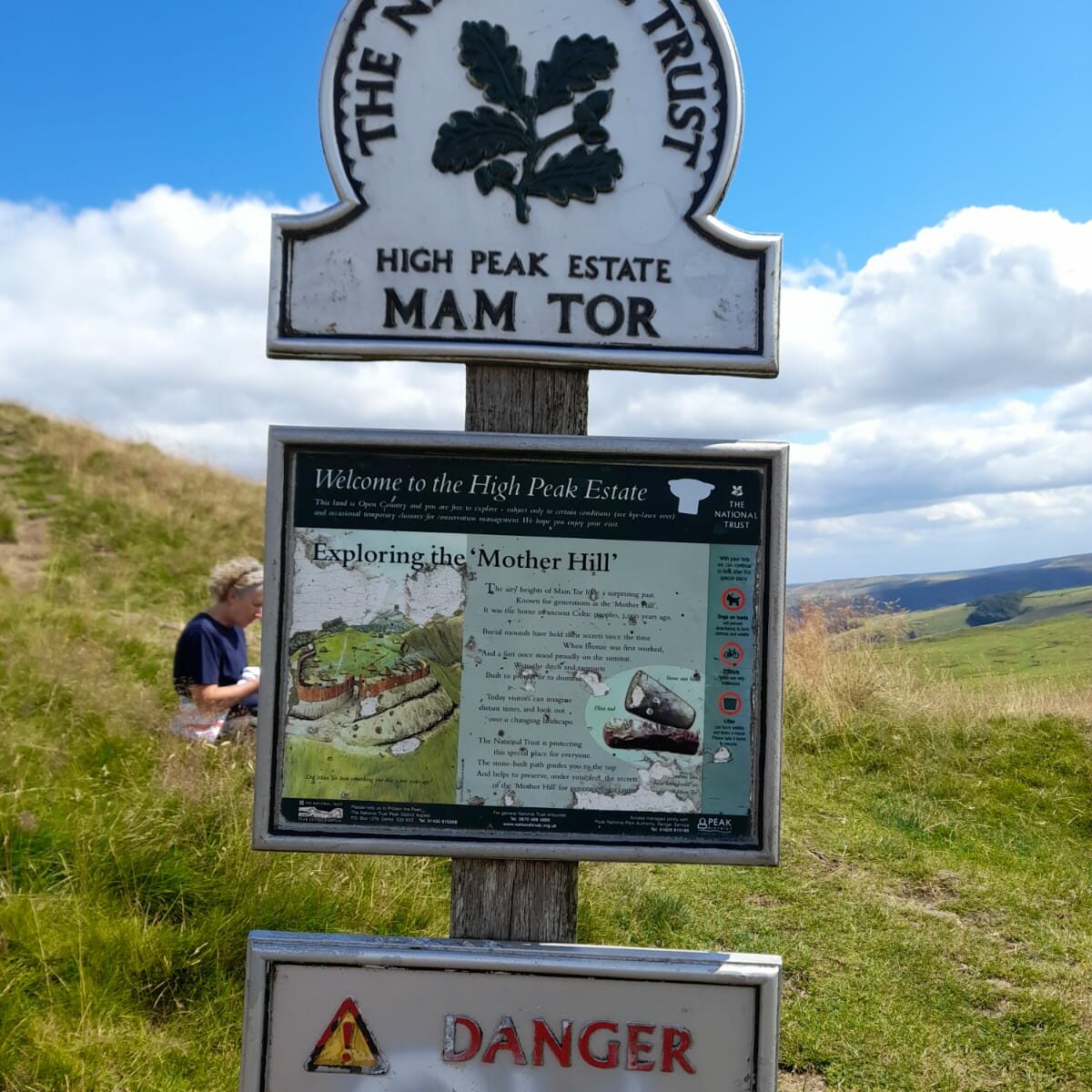 Signpost at Mam Tor, High Peak Estate, with a National Trust logo at the top. Below, there is an information board about Mam Tor, referred to as the 'Mother Hill,' including details about its geology and history. A warning sign beneath it advises visitors to be cautious of cliff edges and to keep children under close supervision. In the background, grassy hills and blue skies are visible, with a person seated in the grass.