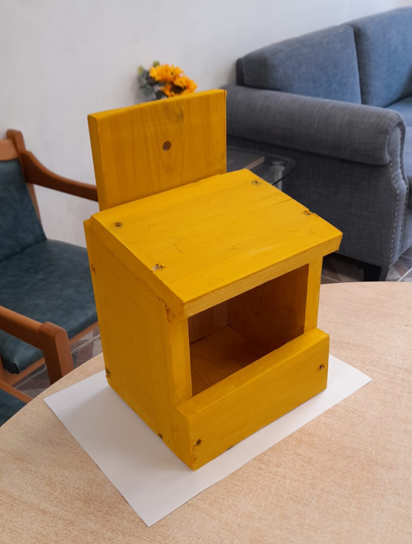 A small wooden birdbox painted in a bright yellow color sits on a table in a cozy living room setting. The birdbox has a simple design with an open front and a flat roof, showcasing visible screws at the corners. In the background, there's a grey sofa and a small bouquet of yellow flowers, adding a homely touch to the scene.