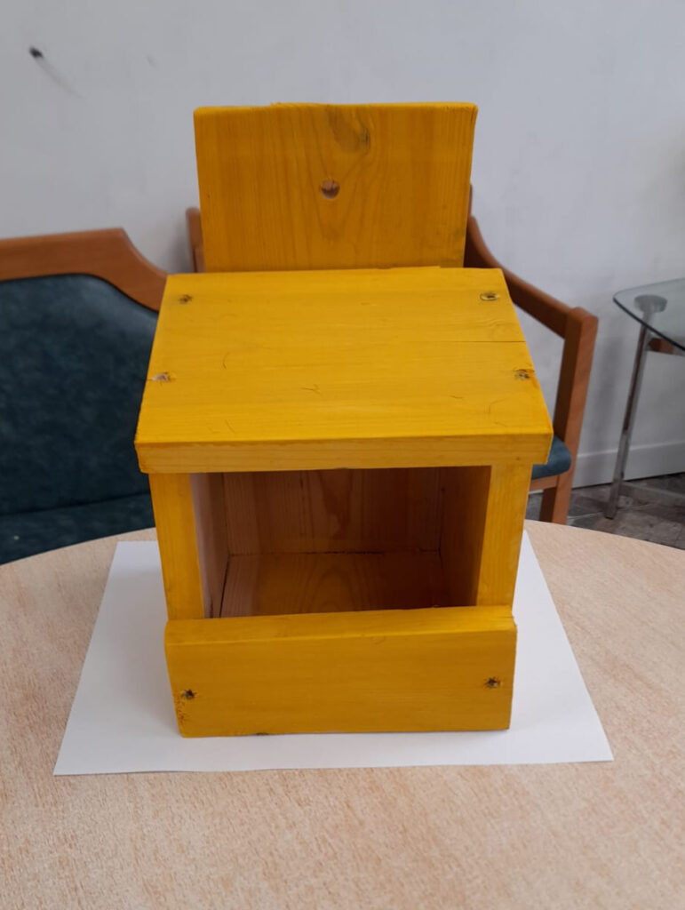 A small wooden birdbox painted in a bright yellow color sits on a table in a cozy living room setting. The birdbox has a simple design with an open front and a flat roof, showcasing visible screws at the corners. In the background, there's a grey sofa and a small bouquet of yellow flowers, adding a homely touch to the scene.