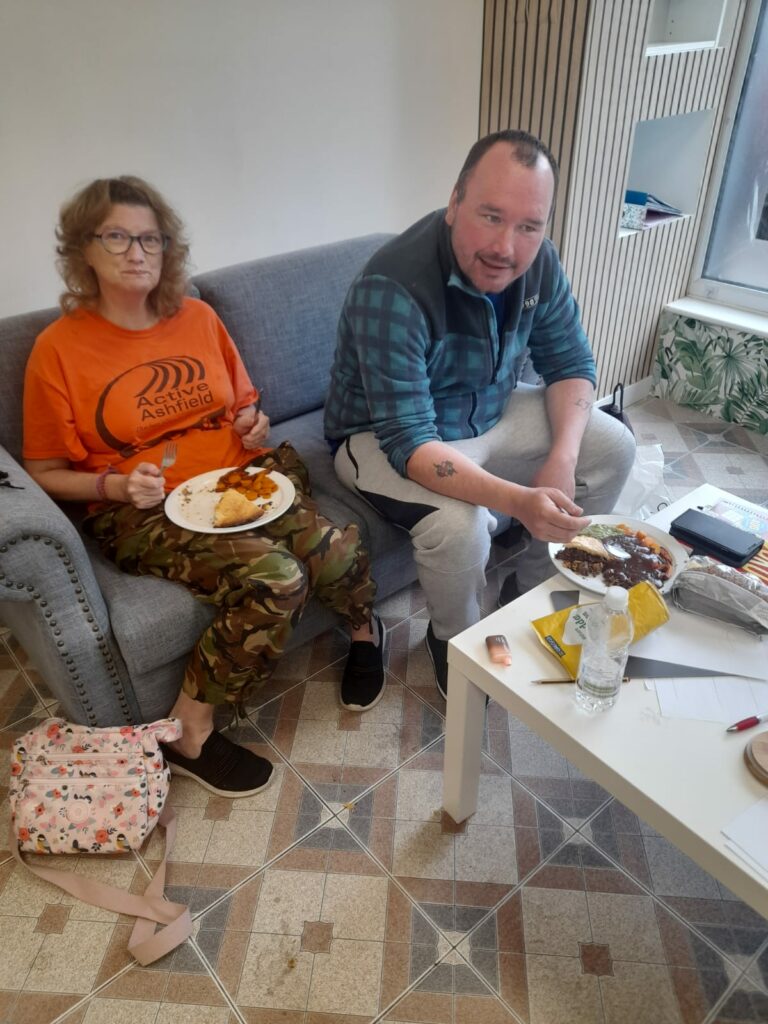 A man and woman sitting on a grey sofa, both holding plates with food. The woman, wearing glasses, an orange 'Active Ashfield' t-shirt, and camouflage trousers, has a plate of sweet potato fries. The man is wearing a checkered fleece jacket and grey sweatpants, holding a fork and plate with a meal covered in gravy. A floral pink bag rests on the floor next to the woman. In front of them is a white coffee table with a water bottle and various items.