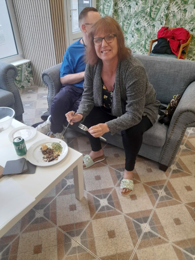 A smiling woman sitting on a grey sofa, holding a fork and plate with a partially eaten meal in her hand. She is wearing glasses, a grey cardigan, and floral sandals. On the white coffee table in front of her, there is a can of Sprite and some scattered papers. Another person is seated next to her, slightly out of view, wearing a blue shirt.