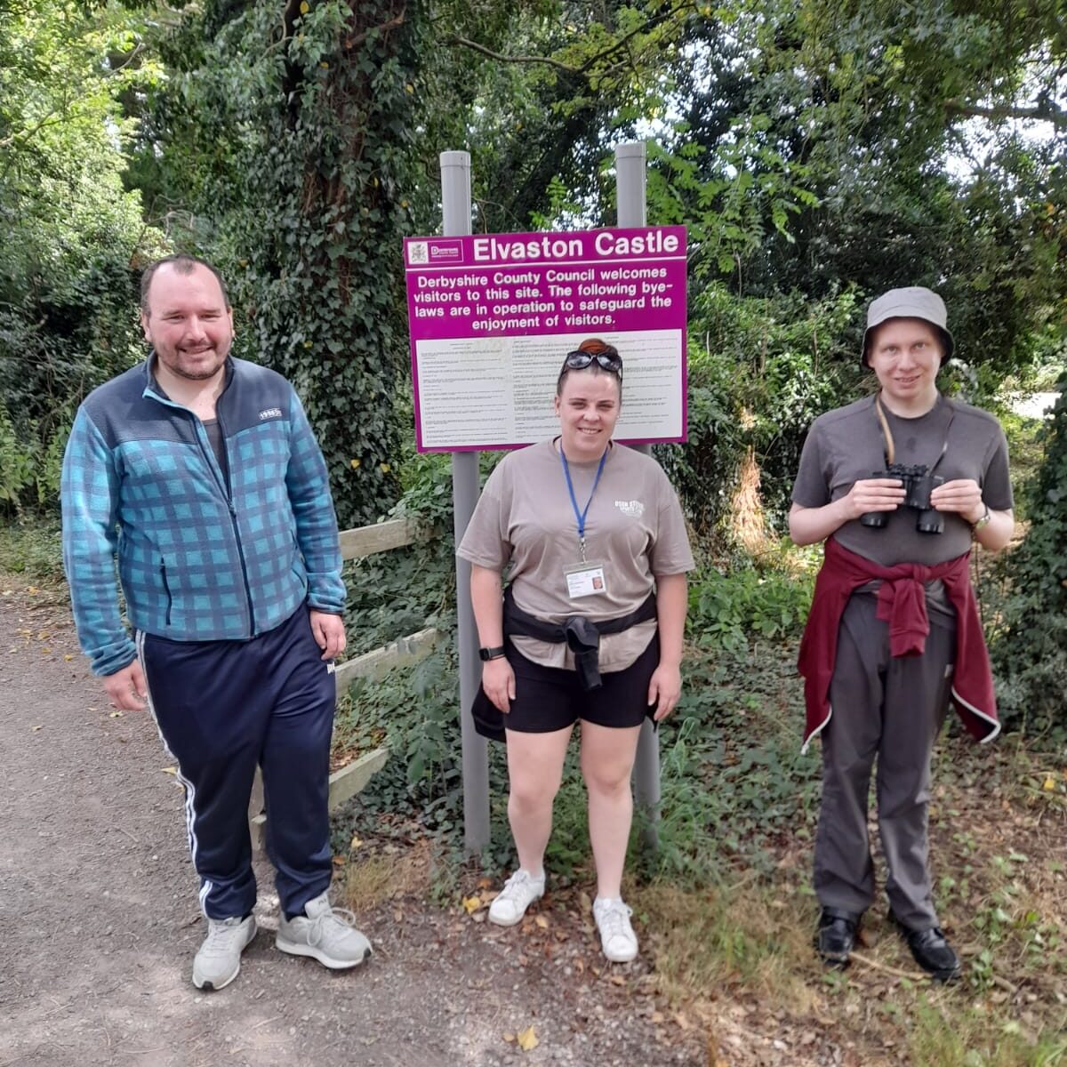 Three individuals stand in front of a sign for Elvaston Castle, which is surrounded by greenery. The person on the left is wearing a blue jacket, the person in the middle is wearing a beige shirt and black shorts, and the person on the right is holding binoculars and wearing a grey t-shirt and hat.