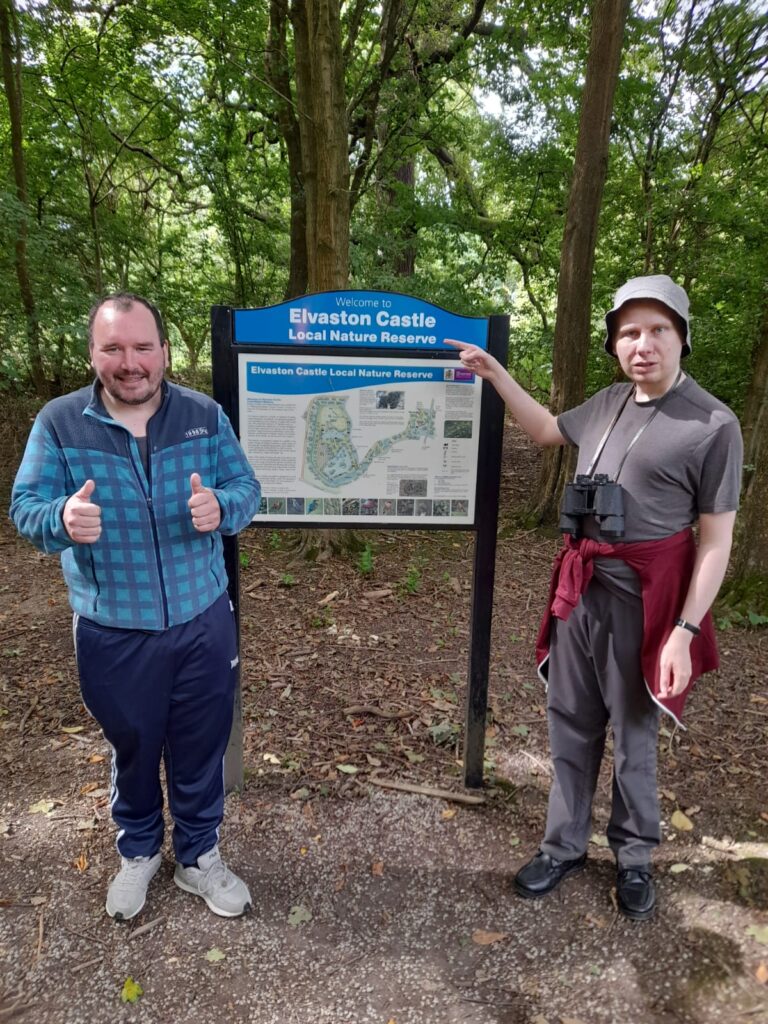 Two people stand by a sign welcoming visitors to Elvaston Castle Local Nature Reserve. One person on the left is giving a thumbs-up, while the person on the right points to the sign and holds binoculars.