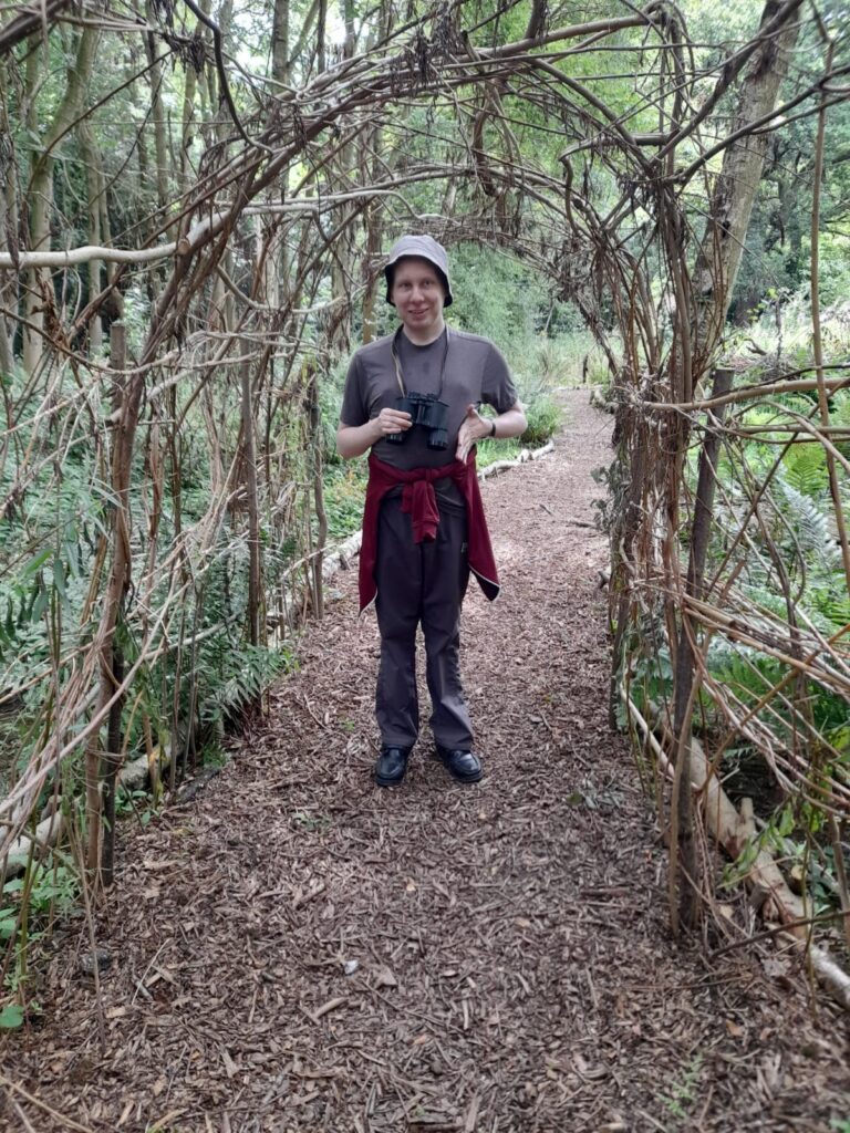 A person wearing a grey t-shirt and bucket hat stands under an archway made from intertwined branches, holding binoculars, in a wooded path.