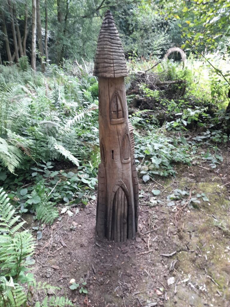 A tall, wooden sculpture resembling a tower or castle spire is set among lush greenery and ferns in a woodland area.