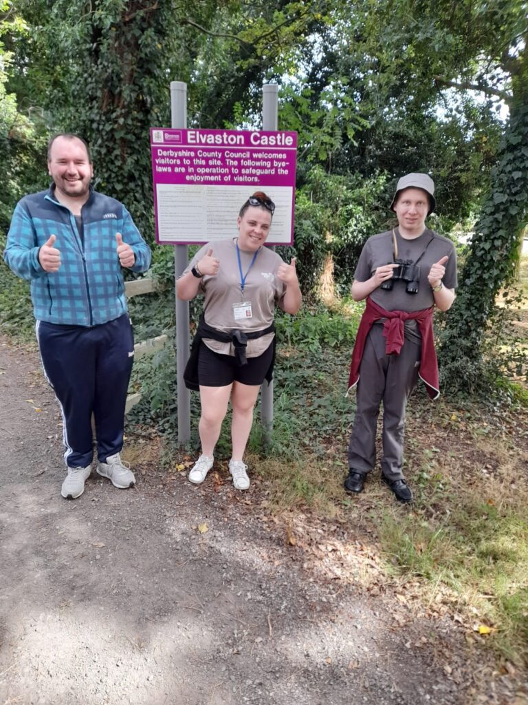 Three individuals give a thumbs-up in front of the Elvaston Castle sign. The person on the left is in a blue jacket, the person in the middle wears a beige shirt and black shorts, and the person on the right has binoculars around their neck and a red jacket tied around their waist.