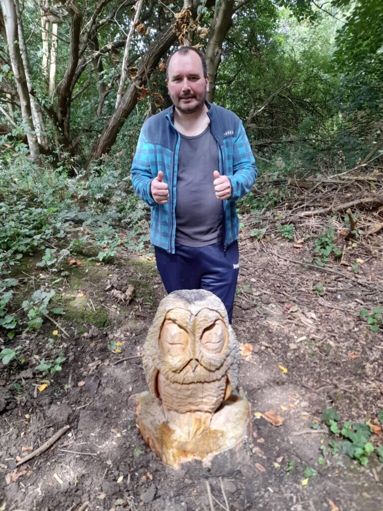 A man in a blue checkered jacket stands outdoors next to a detailed owl carving made from wood. The background features trees and undergrowth, creating a woodland atmosphere.