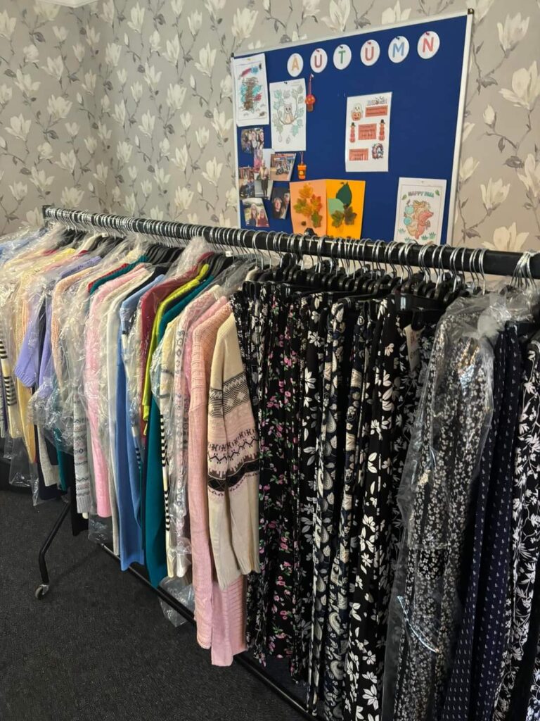 A clothing rack lined with various colorful and patterned clothing items, in front of an autumn-themed notice board displaying art and seasonal decorations.