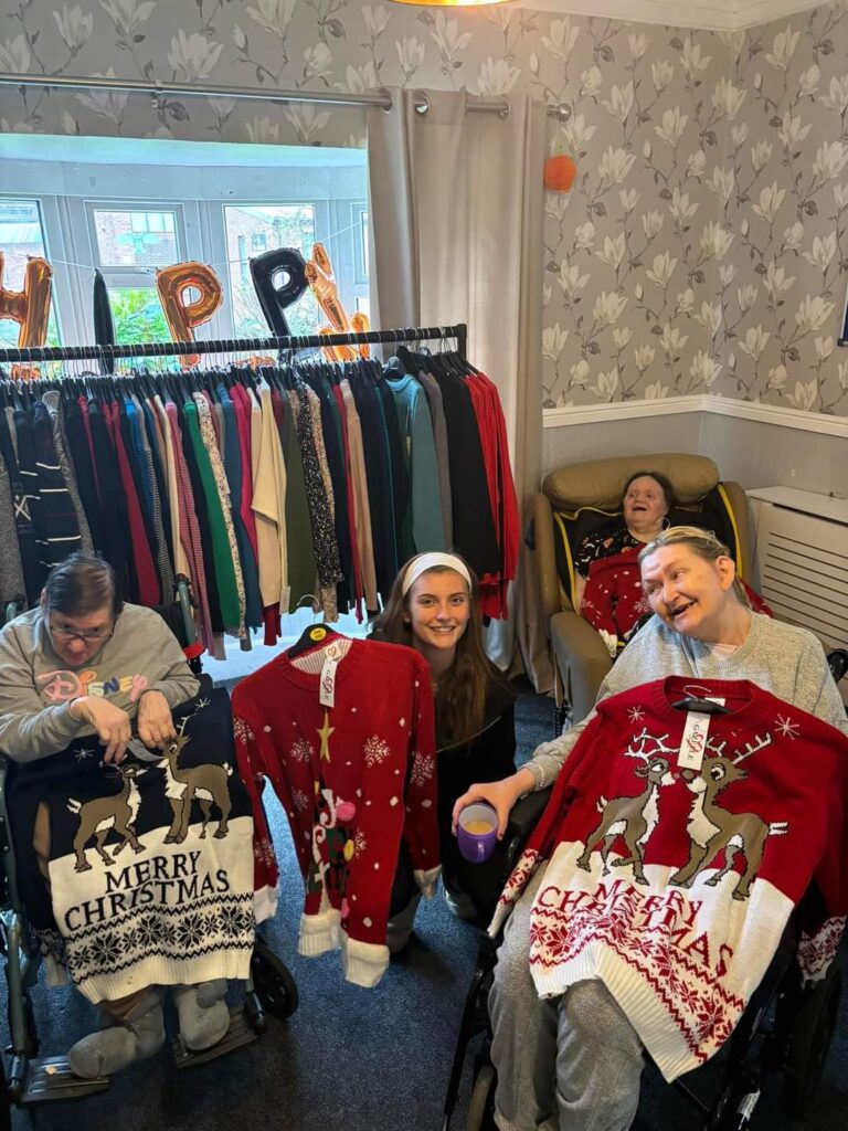 A group of residents smiling and showing off festive Christmas jumpers in front of clothing racks. The room has a celebratory atmosphere with "HAPPY" letter balloons in the background, and everyone is engaged in the festive spirit.