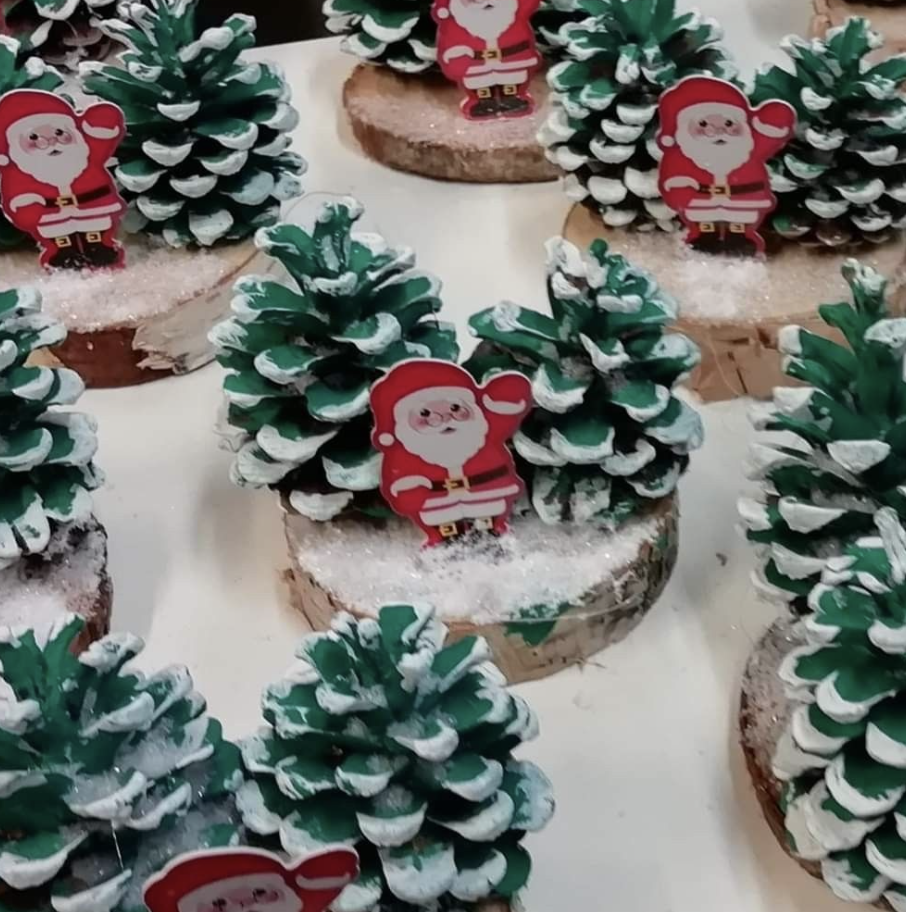 Handcrafted Christmas decorations featuring painted pinecones with a dusting of artificial snow, mounted on wooden slices. Small cutout images of Santa Claus waving are placed beside each pinecone, creating a festive holiday display.