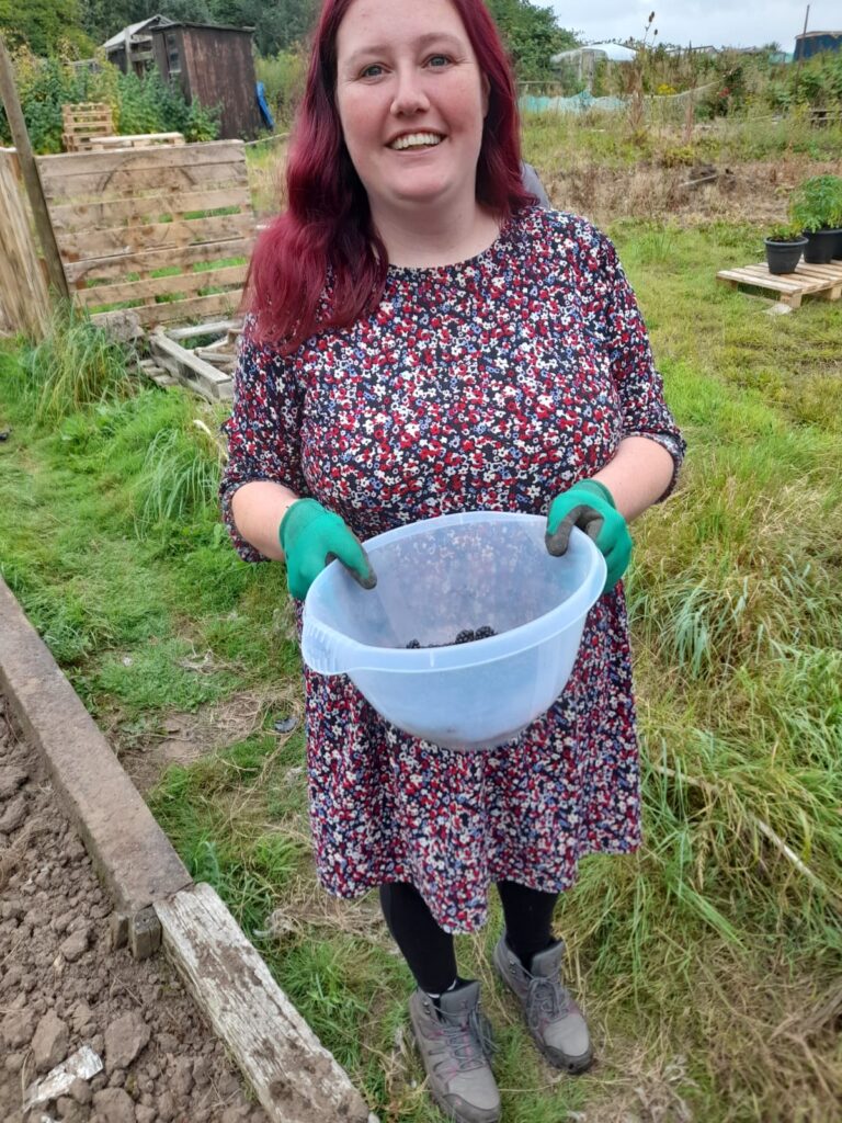 Woman Smiling with Harvest: A woman with red hair smiles while holding a plastic bowl containing freshly picked blackberries. She is wearing a floral dress, green gloves, and sturdy boots, standing in a grassy area near an allotment.