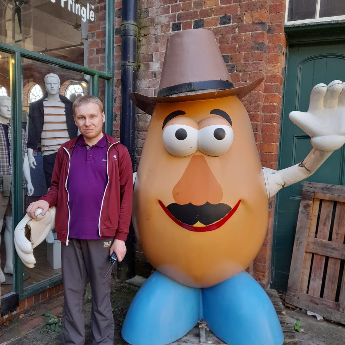 A man in a burgundy jacket and purple polo shirt standing next to a large, smiling potato statue wearing a cowboy hat, outside a James Pringle store.