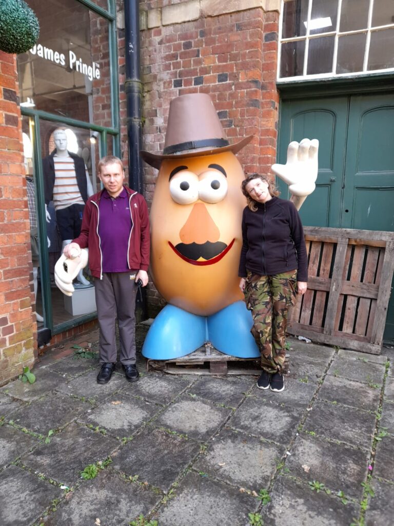 A man in a burgundy jacket and purple polo shirt stands next to a large potato statue wearing a cowboy hat, with a woman dressed in camouflage trousers leaning her head on the statue's arm, both smiling outside a James Pringle store.