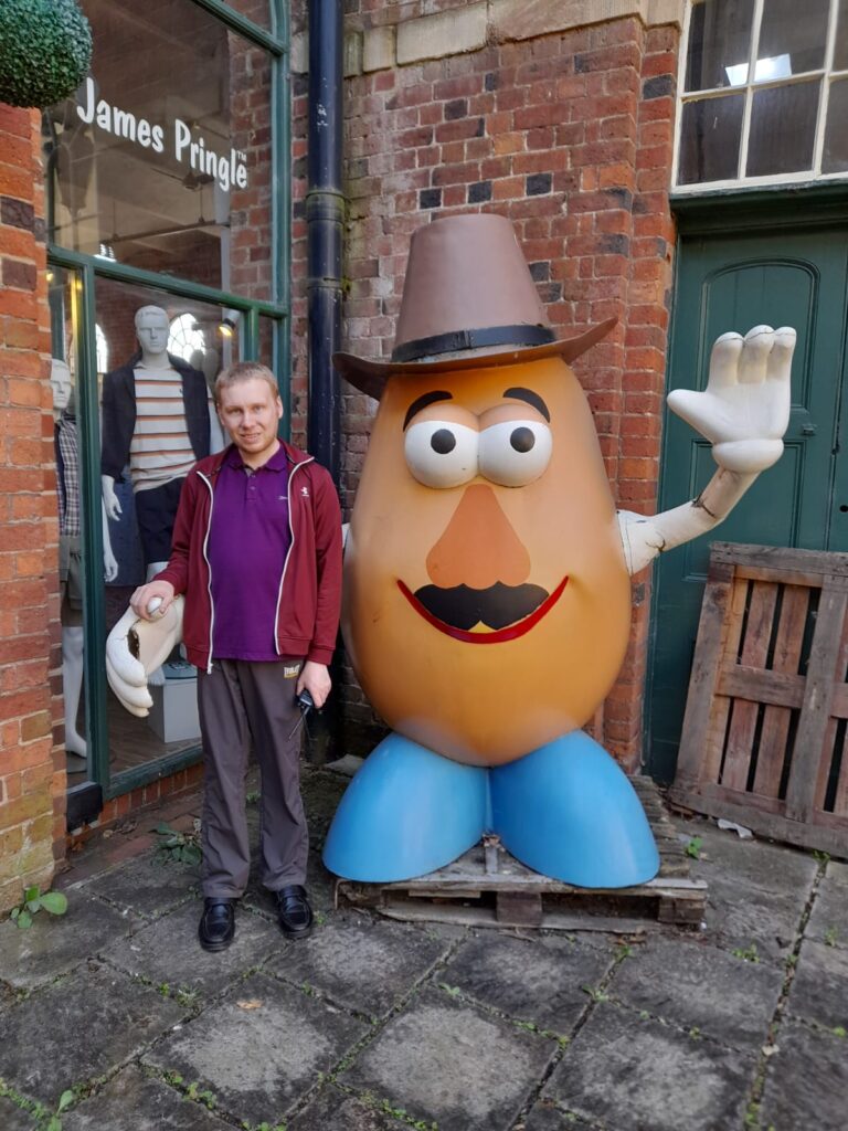 A man in a burgundy jacket and purple polo shirt standing next to a large potato statue wearing a cowboy hat, raising one hand as if to wave, outside a James Pringle store.