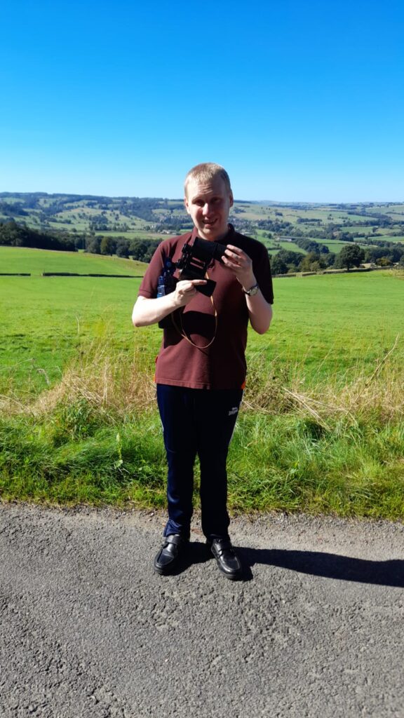 Man with binoculars: A man stands alone on the road holding binoculars, examining them carefully. He is wearing a brown shirt and dark pants, standing against a scenic backdrop of green fields and distant hills under a bright blue sky.