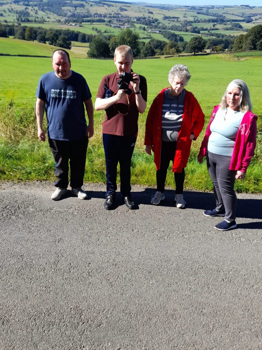 Group Photo with Scenic Background: Four people standing side by side on a road with a picturesque rural landscape in the background. Rolling green fields stretch into the distance under a clear blue sky. From left to right, a man in a navy blue T-shirt, a man in a brown shirt holding a camera, an elderly woman in a red jacket, and a woman with long grey hair in a light blue top and pink hoodie.