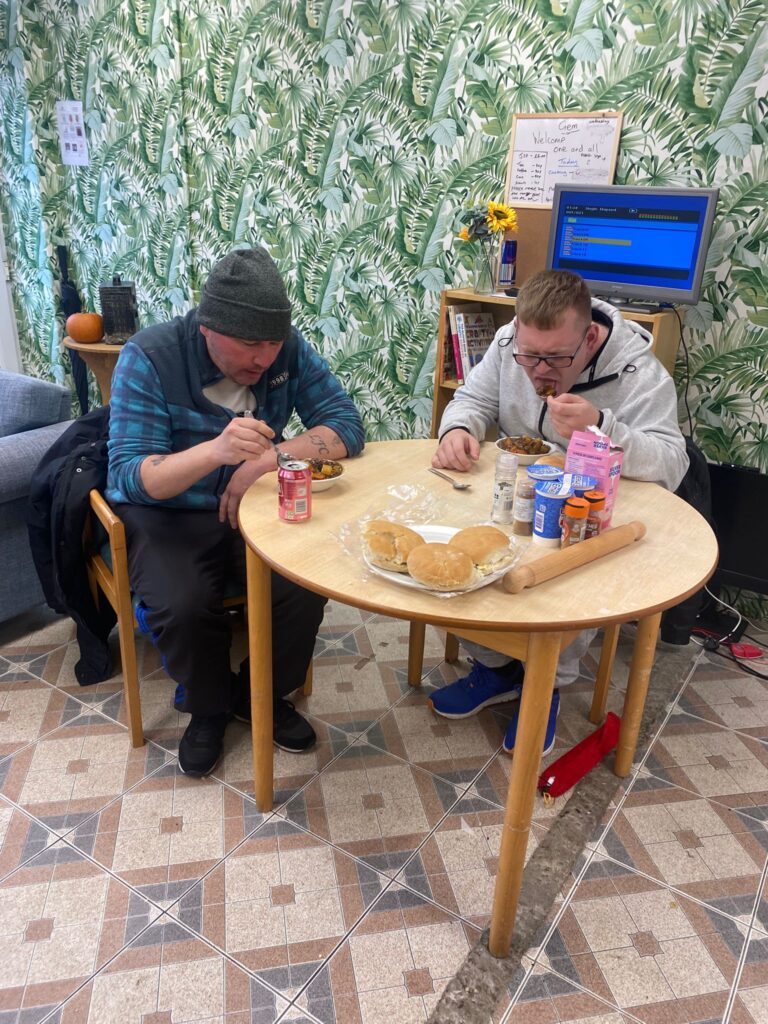 Two men sitting at a round table eating from bowls. They are engaged in conversation while eating, with buns and condiments on the table. The room has green leafy wallpaper, a tiled floor, and a small monitor in the background.