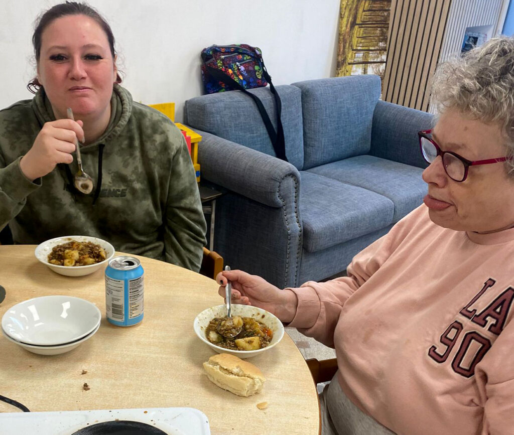 Two women sitting at a table, enjoying bowls of stew. One woman is wearing a green hoodie, and the other an LA-themed pink sweatshirt. There is a bread roll and cans of soda on the table in front of them, suggesting they are having a meal together.