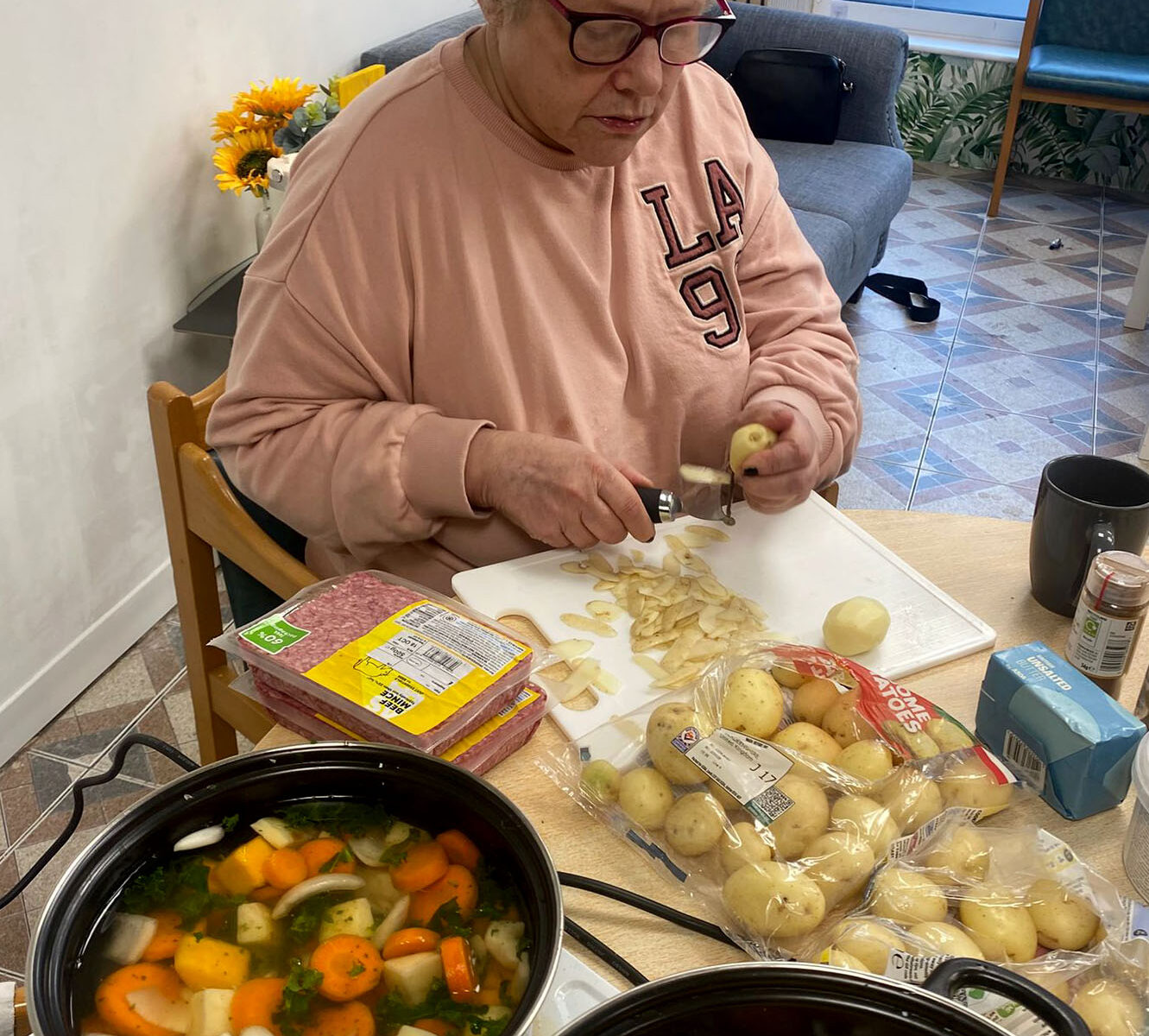 An older woman peeling potatoes at a table, surrounded by ingredients including carrots and potatoes. Two large pots of vegetable soup or stew are cooking on the table in front of her.