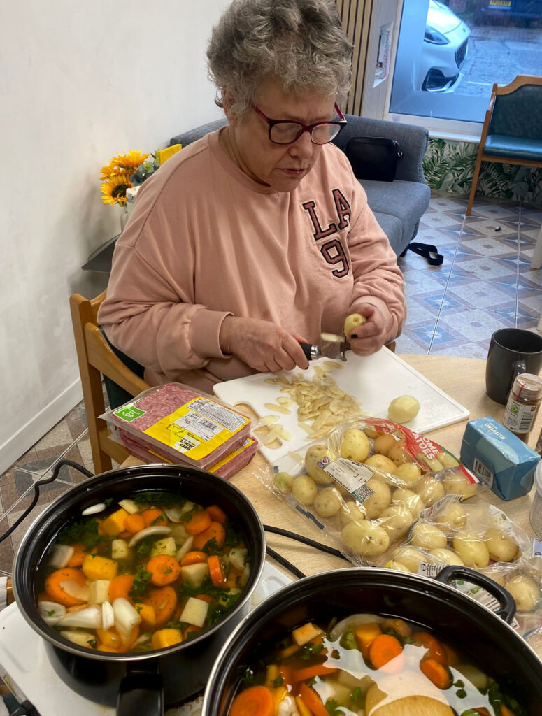 An older woman peeling potatoes at a table, surrounded by ingredients including carrots and potatoes. Two large pots of vegetable soup or stew are cooking on the table in front of her.