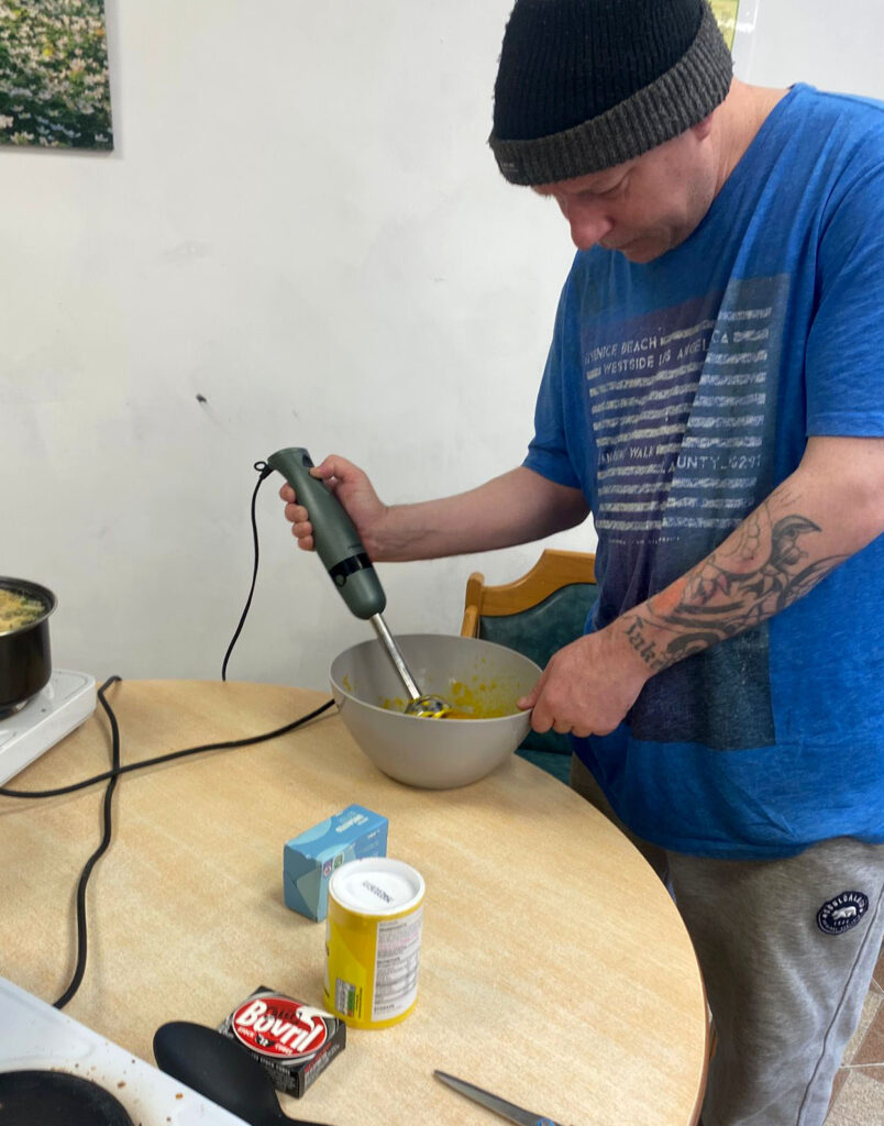 A man wearing a beanie and a blue shirt is using an immersion blender to mix ingredients in a large bowl on a table. Cooking supplies and food items are placed nearby, with a pot of soup in the background.