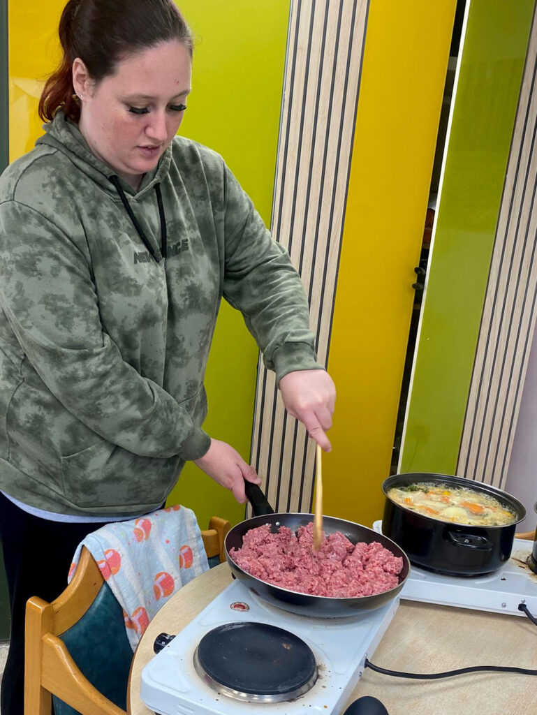 A woman wearing a green hoodie is stirring minced meat in a frying pan on a portable stove. Another pot of soup or stew is simmering beside her on the table, which is also part of a cooking session.