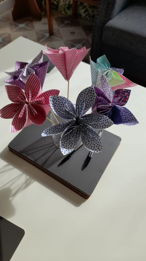 A collection of vibrant origami paper flowers made at the community hub, displayed in a small vase. The flowers are crafted from various patterned papers, including stripes, hearts, and polka dots, in colors like red, purple, blue, and green. They are arranged neatly on a white surface, with a soft shadow cast by the sunlight.