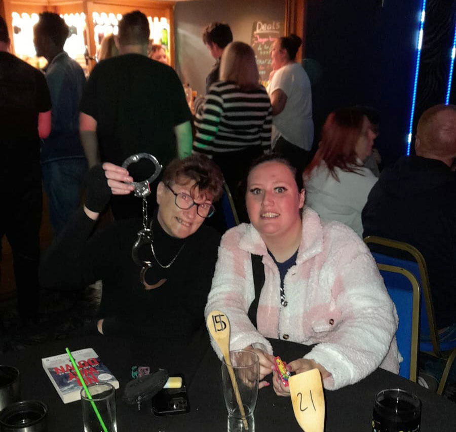 Two women seated at a table at The Towers Disco. One is holding up a pair of toy handcuffs and smiling, while the other woman, in a white and pink fluffy jacket, smiles beside her. The background shows other attendees near the bar area, enjoying the event.