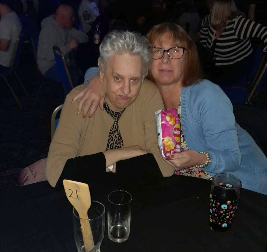 Two women sitting closely together at The Towers Disco. One woman with short grey hair and a beige cardigan has a serious expression, while the other, wearing glasses and a blue cardigan, hugs her with a smile and holds a bag of crisps. Empty glasses and table markers are visible in the foreground.