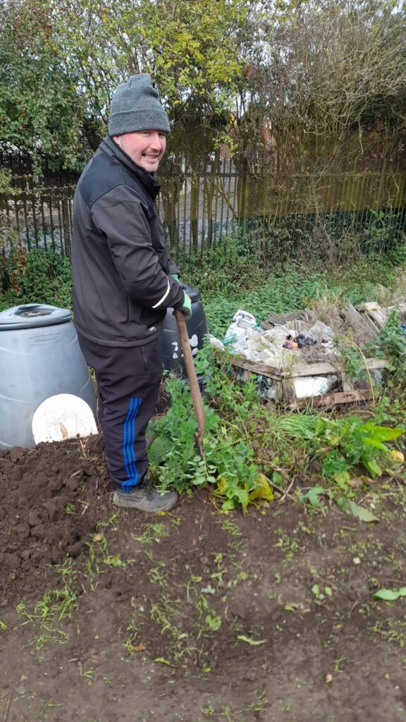 A man wearing a grey beanie and black jacket, working at the allotment. He is smiling while holding a garden fork and standing near some compost bins and weeds.