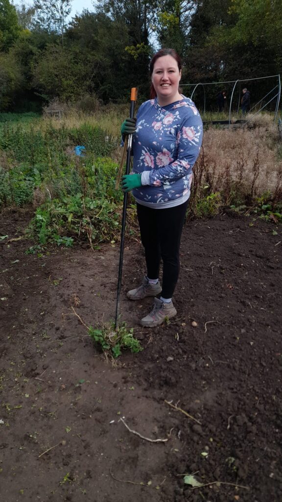 A woman wearing a floral hoodie and gloves, standing in a garden bed at the allotment. She is smiling while holding a garden fork, with the surrounding area showing some cleared soil and greenery.