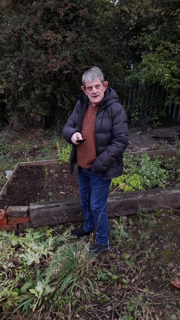 A man standing in front of a raised garden bed at the allotment. He is dressed warmly in a black jacket and brown jumper, holding a small tool, with the surrounding area covered in greenery.