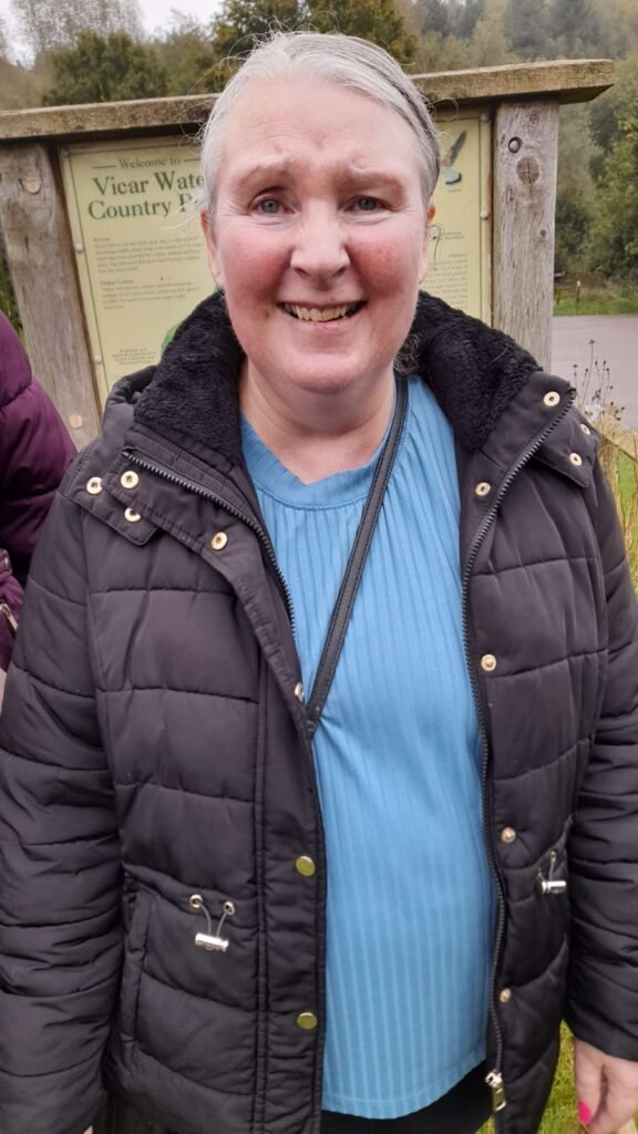 A smiling woman in a black puffer jacket standing in front of a sign at Vicar Water Country Park. She looks happy and ready for the walk, with trees and greenery in the background.