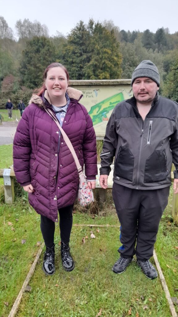 Two people standing together, both smiling, in front of the Vicar Water Country Park sign. The woman is wearing a purple winter coat and the man is dressed in a grey beanie and black jacket. Behind them, trees and other walkers can be seen.