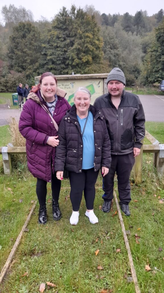 Three people standing together, smiling, in front of the Vicar Water Country Park sign. The woman in the center wears a black puffer jacket, while the other woman is in a purple coat, and the man is in a black jacket and grey beanie. The background shows the park’s green landscape.