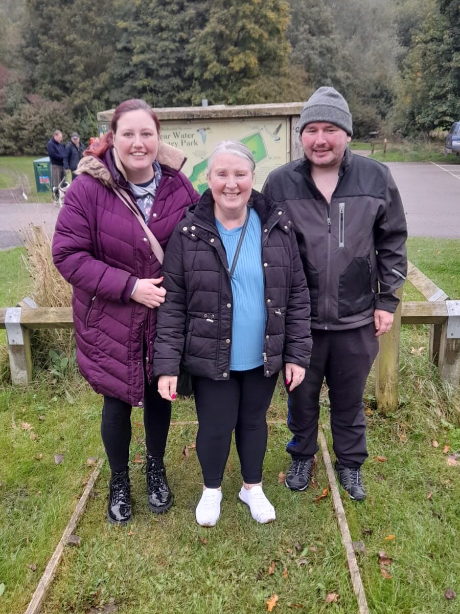 Three people standing together, smiling, in front of the Vicar Water Country Park sign. The woman in the center wears a black puffer jacket, while the other woman is in a purple coat, and the man is in a black jacket and grey beanie. The background shows the park’s green landscape.