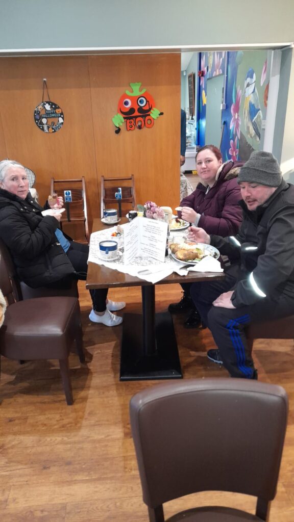 A group of three people sitting at a table in Rumbles Café, enjoying a meal. The table is filled with plates of food, and a pumpkin Halloween decoration is hanging on the wall behind them.