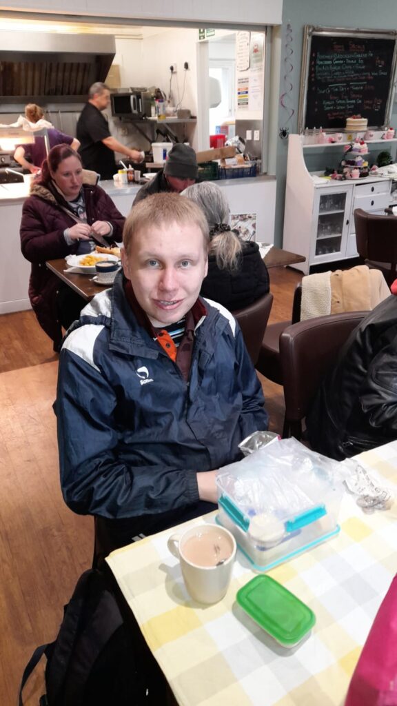 A man sitting at a table inside Rumbles Café, smiling at the camera. He has a packed lunch in front of him, along with a cup of tea, while others in the background are also enjoying their meals.