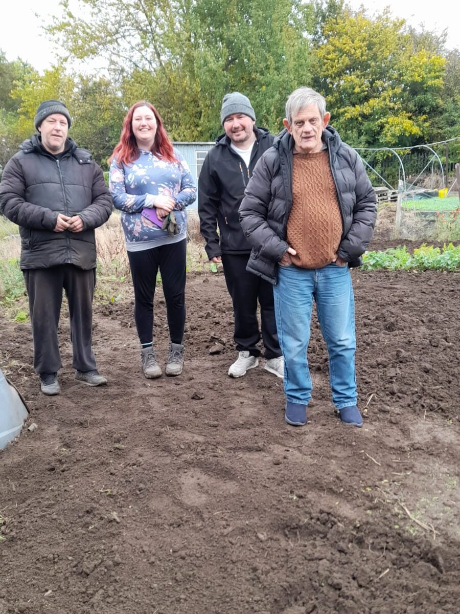 Four gardening club members standing on a tilled section of the allotment, smiling and dressed warmly in outdoor attire.